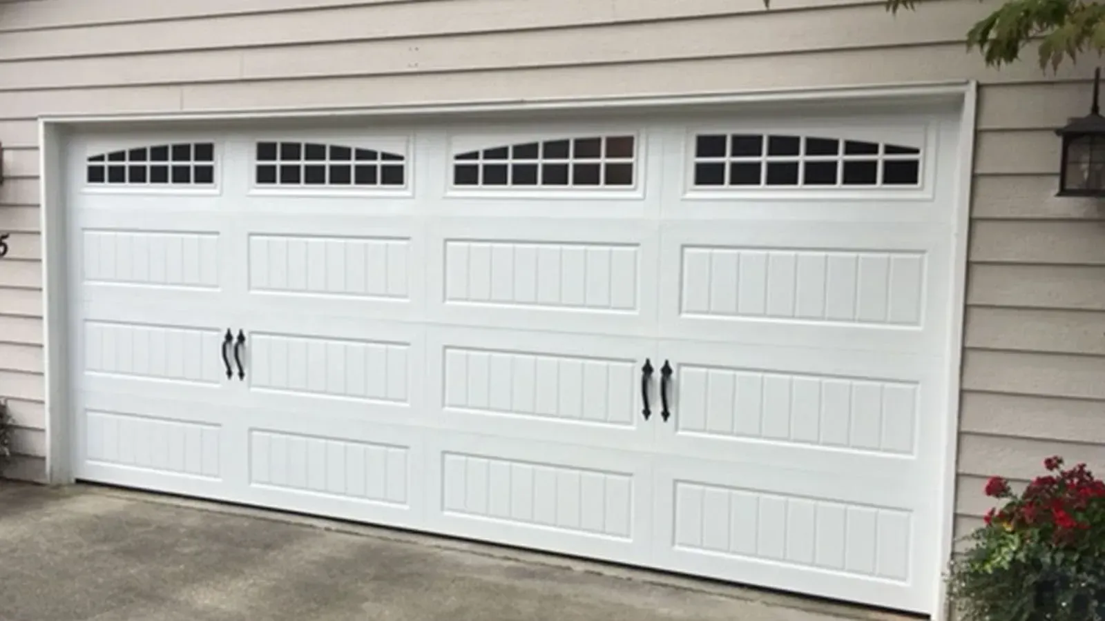 A white two-car garage door with arched windows and two black decorative handles on a home with light-colored siding.