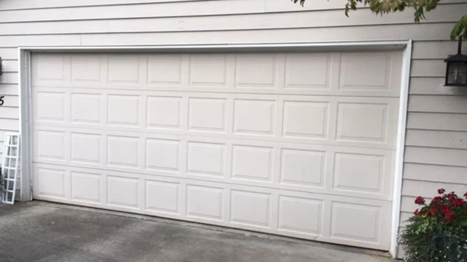 A white, multi-panel garage door on a house with light-colored siding, a small plant, and an outdoor light fixture.