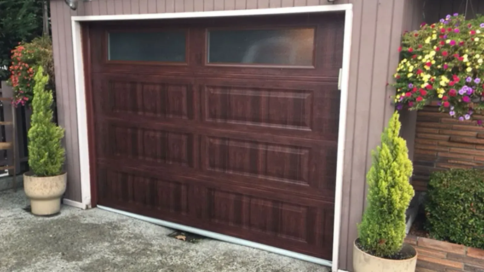 Dark wood garage door with two top glass panels, flanked by potted evergreen trees and hanging flower baskets.