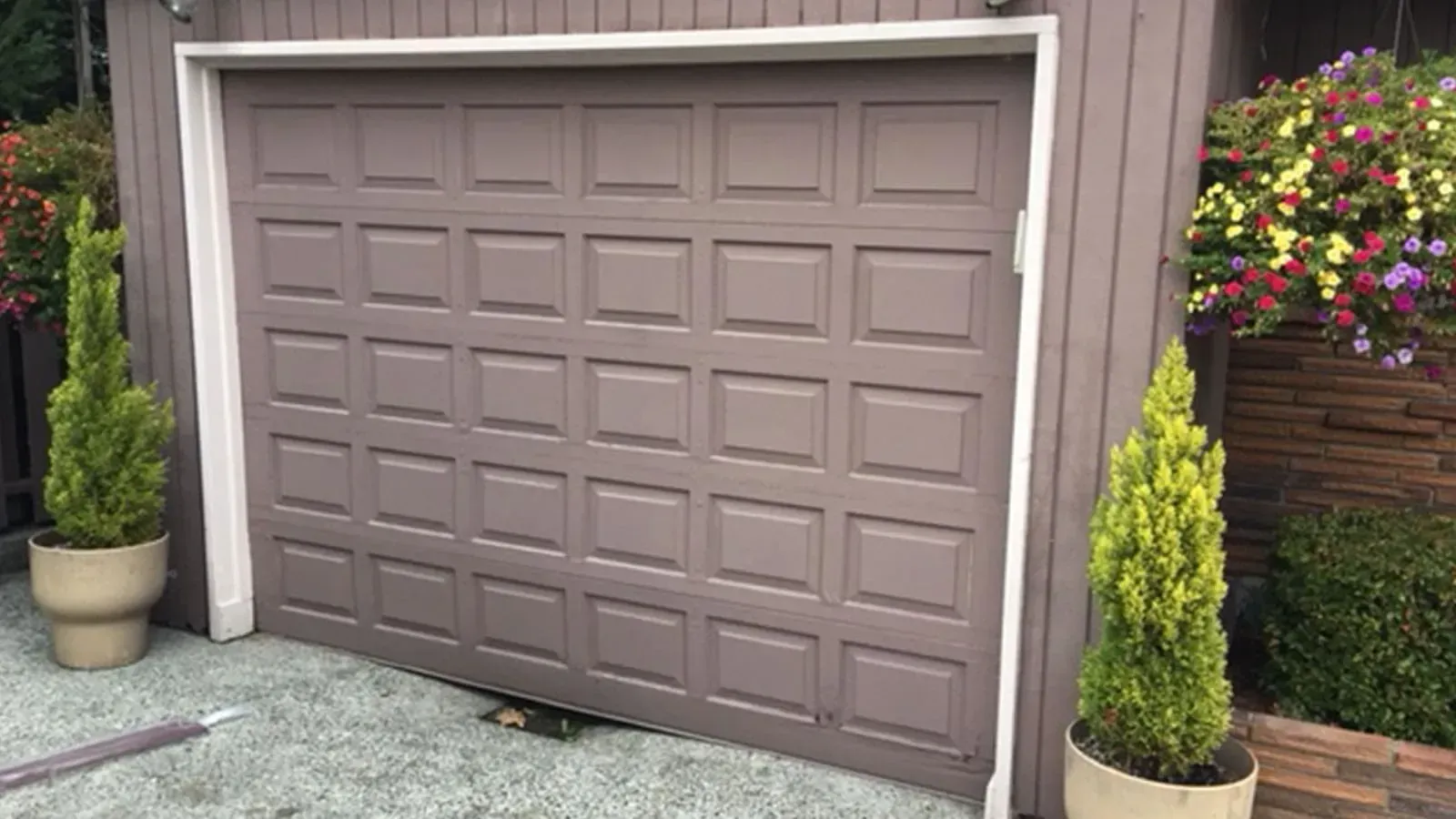 A taupe garage door framed by two potted evergreen trees and hanging flower baskets.