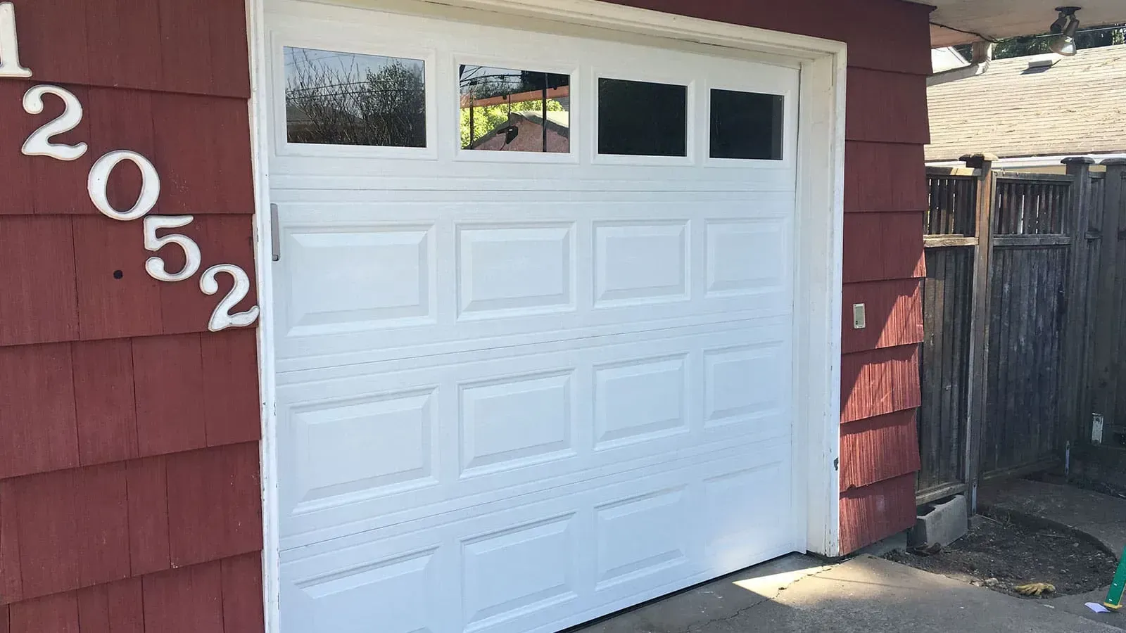 A white garage door with a four-pane window set in a reddish-brown shingled house wall featuring the address number 12052.