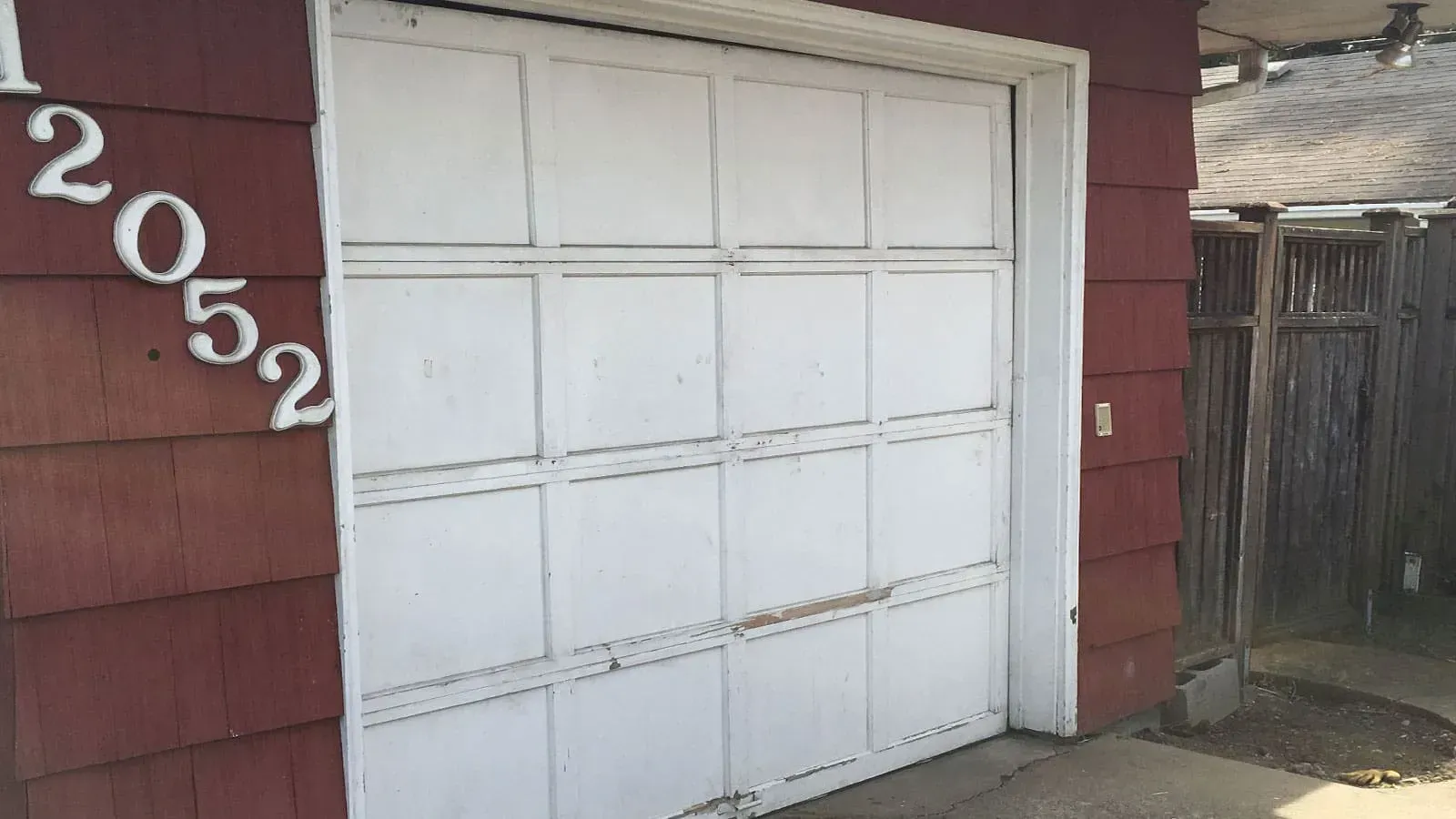A white garage door is set into a red-shingled building with the address number 12052 mounted on the left side.