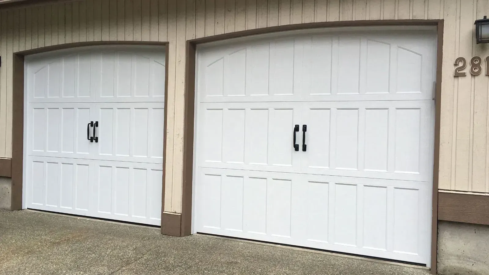 Two white carriage-style garage doors with arched tops and black decorative handles on a light-colored house.