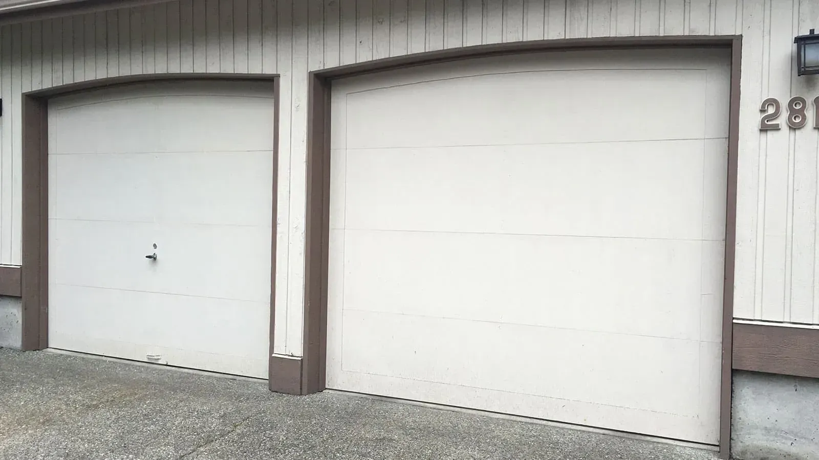 Two off-white, arched-top garage doors on a light-colored, vertical-paneled building with dark brown trim.