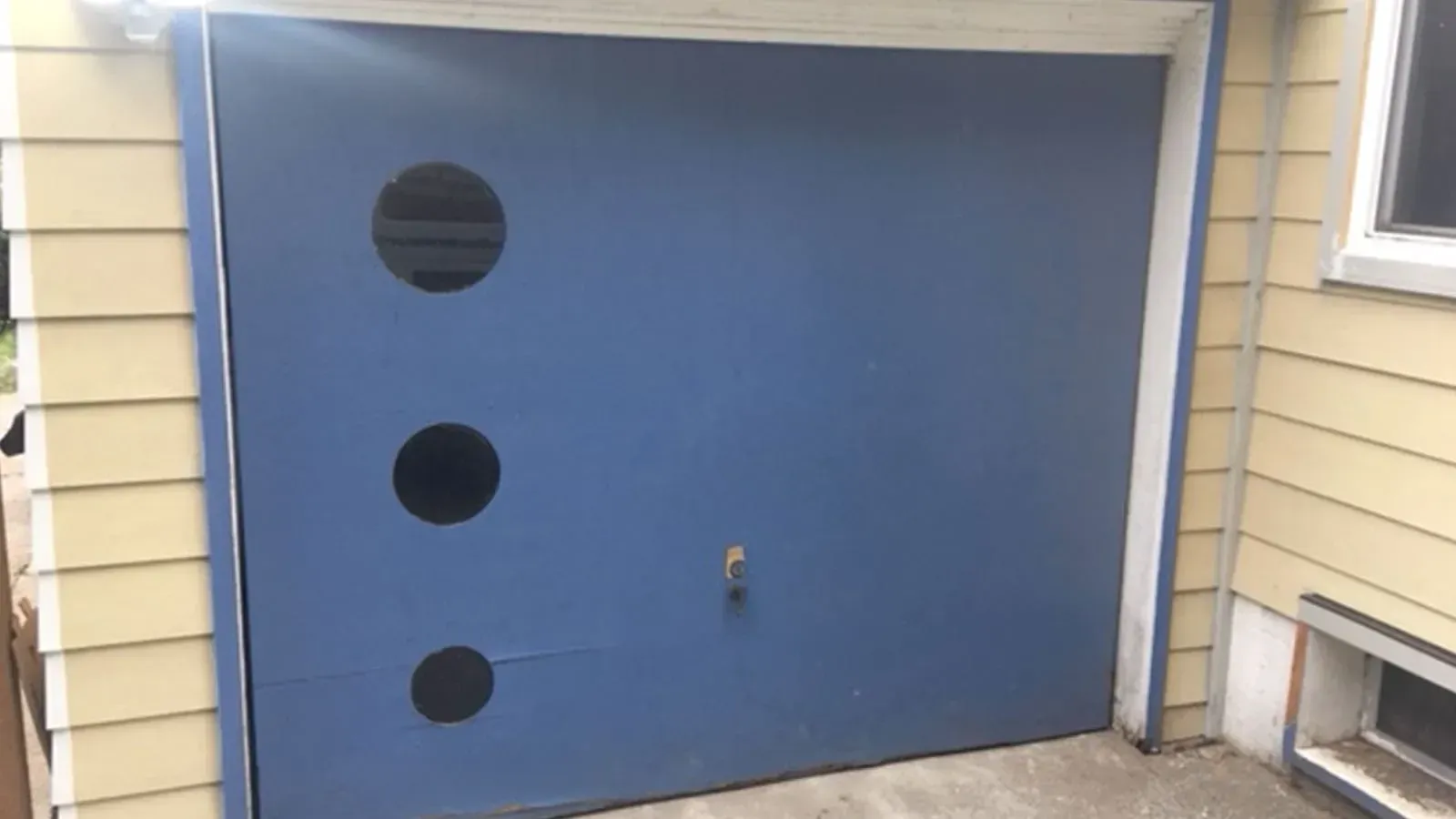 A blue garage door with three round black windows in a vertical line on the left side, set in a light yellow house.