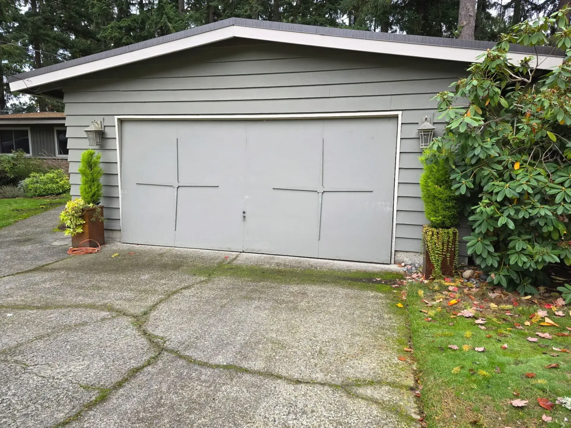 A gray garage with a smooth door and decorative cross-shaped accents, flanked by two potted green shrubs on a driveway.