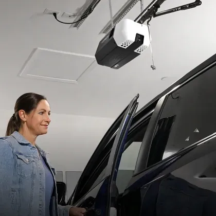 A person stands next to a car with an open door in a garage with a ceiling-mounted garage door opener.