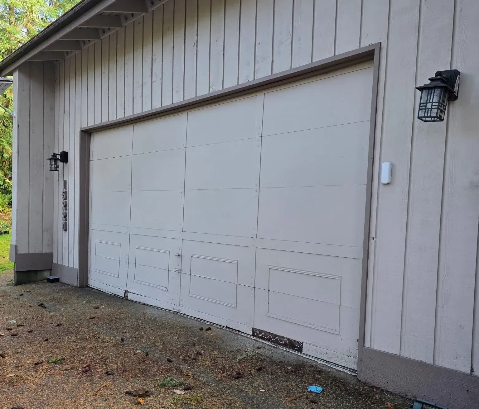 A tan-colored wooden garage exterior with a large, multi-panel garage door, a side wall lamp, and house numbers.