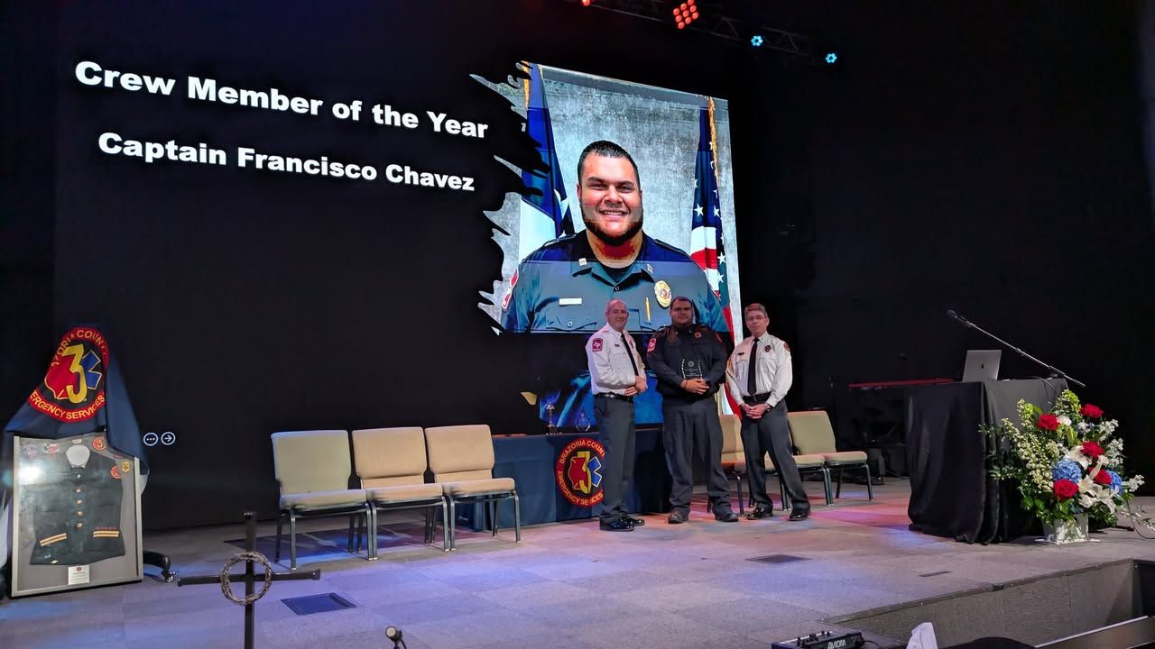Three people on a stage during an awards ceremony, with a portrait of Captain Francisco Chavez on a large screen behind.