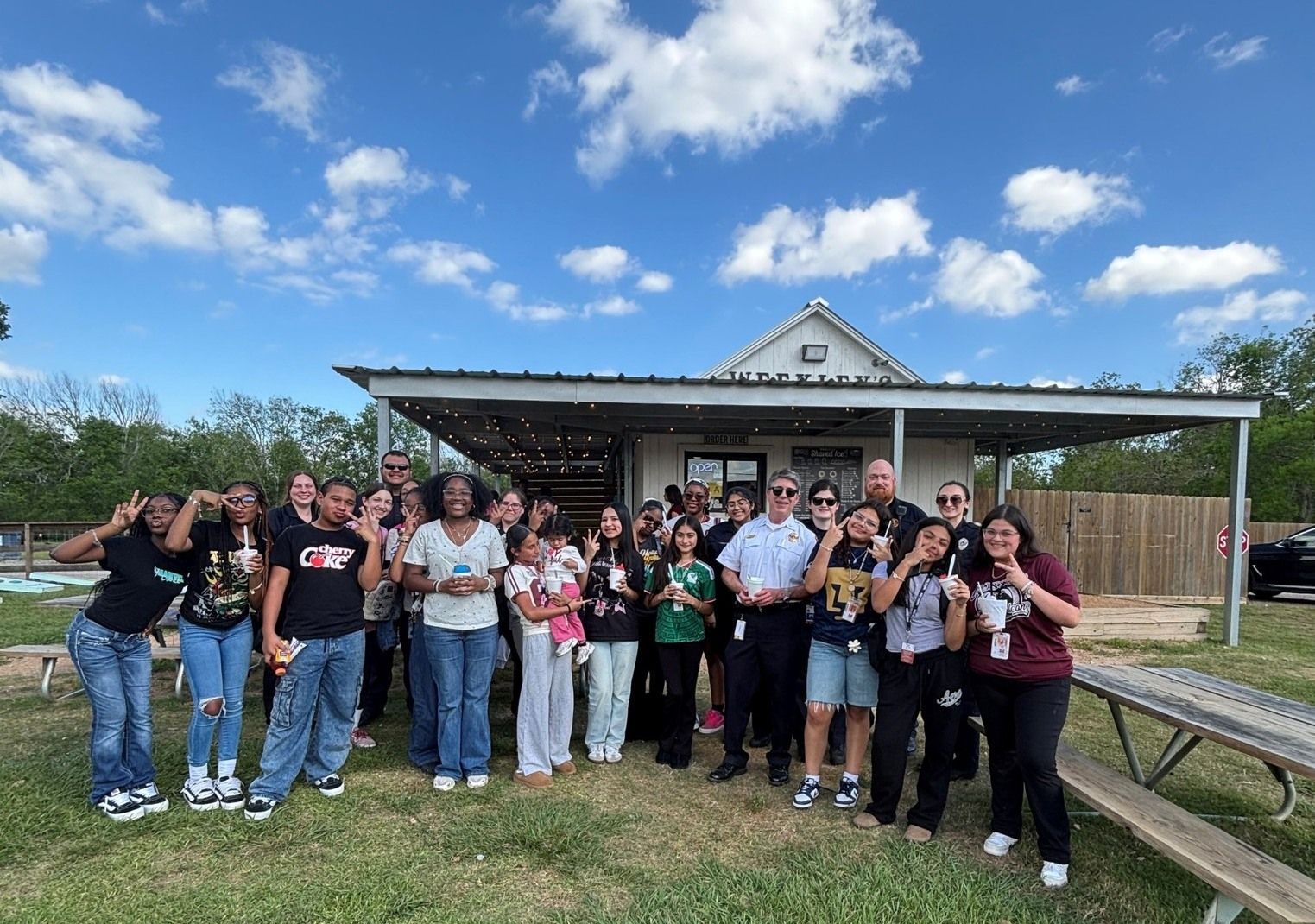 A group of people standing together outdoors in front of a covered patio building under a blue sky.
