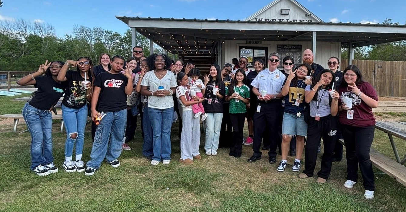 A large group of people smiling and posing for a photo outdoors in front of a covered patio building on a sunny day.
