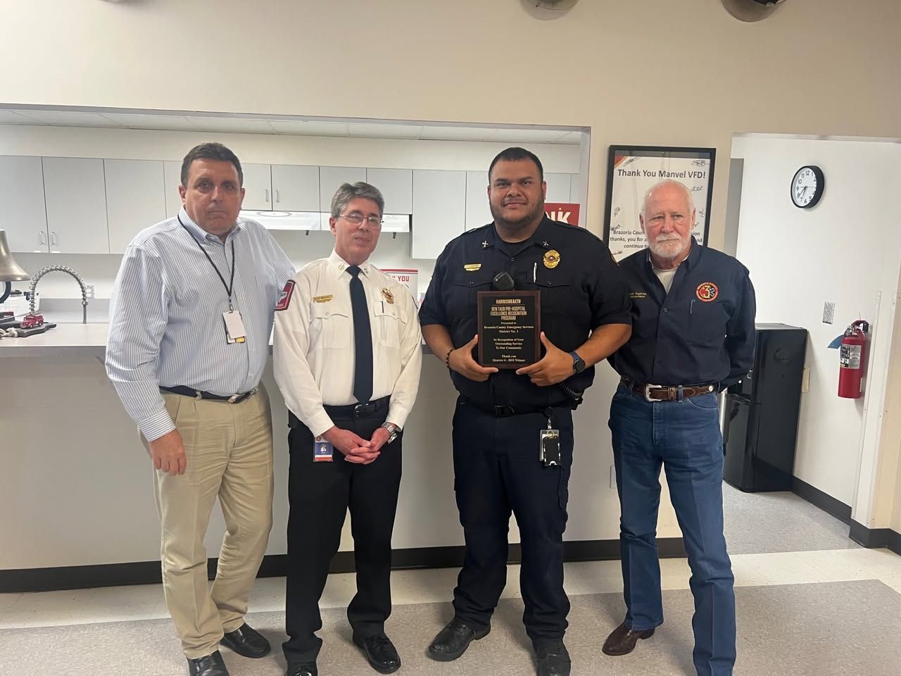 Four men stand indoors; one in a uniform holds an award plaque in front of a white wall and counter.