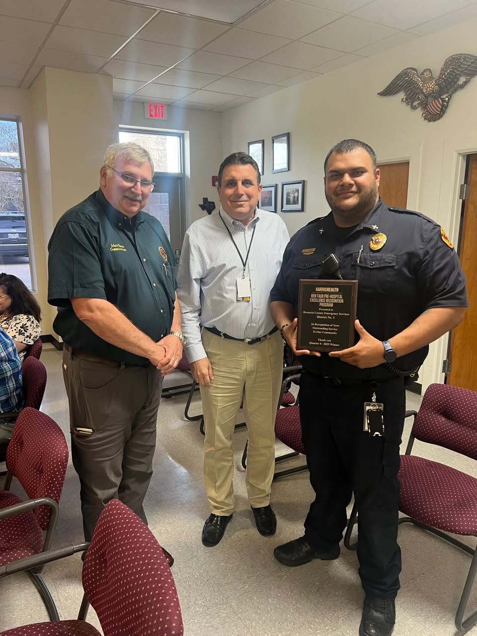Three men standing in an office, one in uniform holding an award plaque, flanked by two others in professional attire.