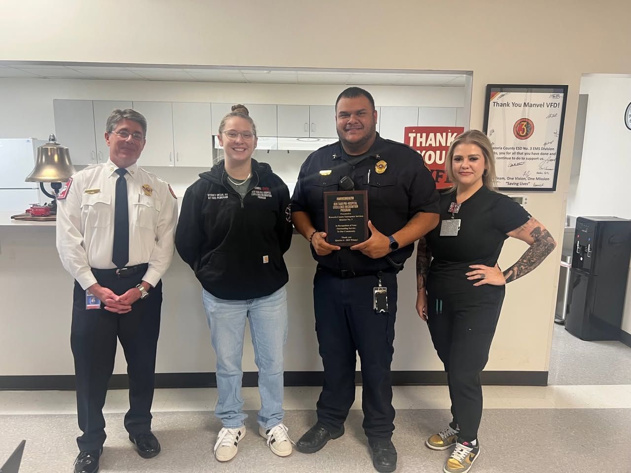 Four people stand in an office; one person in uniform holds a plaque while others pose for a celebratory photo.
