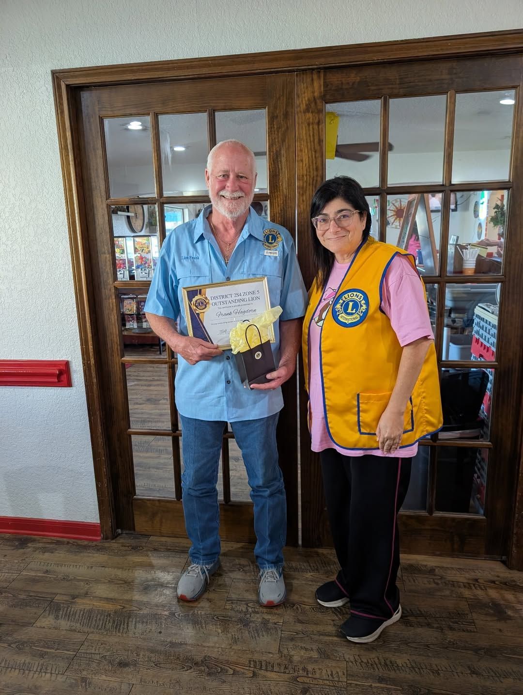Two people pose indoors by glass doors; one holds a framed certificate and the other wears a yellow vest.