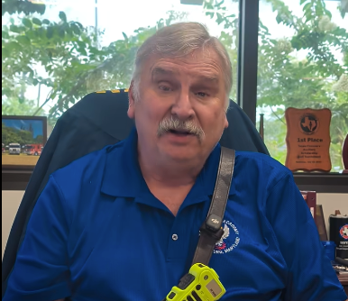 Man in blue polo speaking at a desk, with books and a trophy behind him.