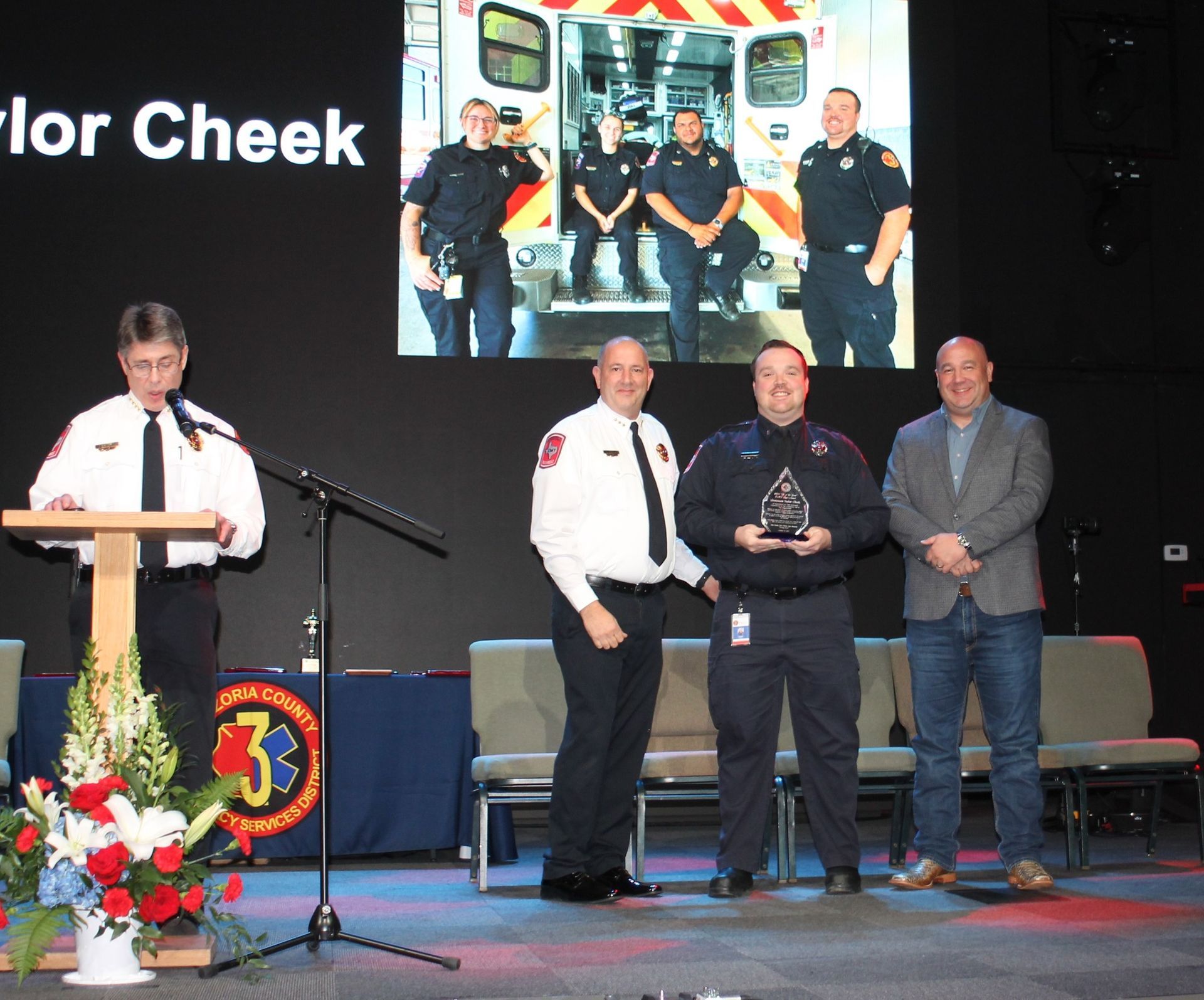 At an awards ceremony, a person at a podium speaks, while two men present an award to another man on stage. A screen in the background shows a group of people in front of an ambulance.
