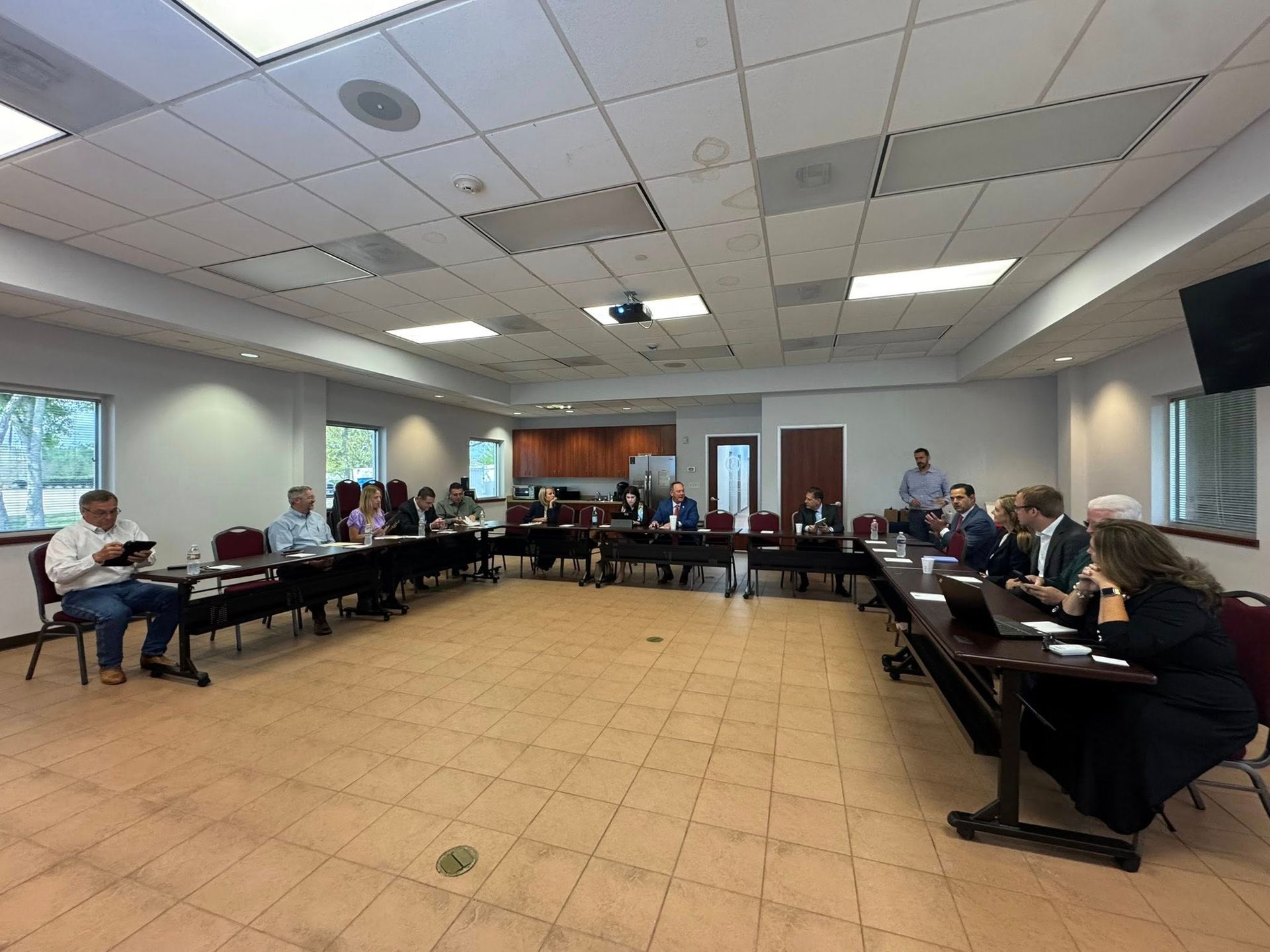 Four people seated at a long table during a meeting in a room with wood cabinets and a stainless steel refrigerator.