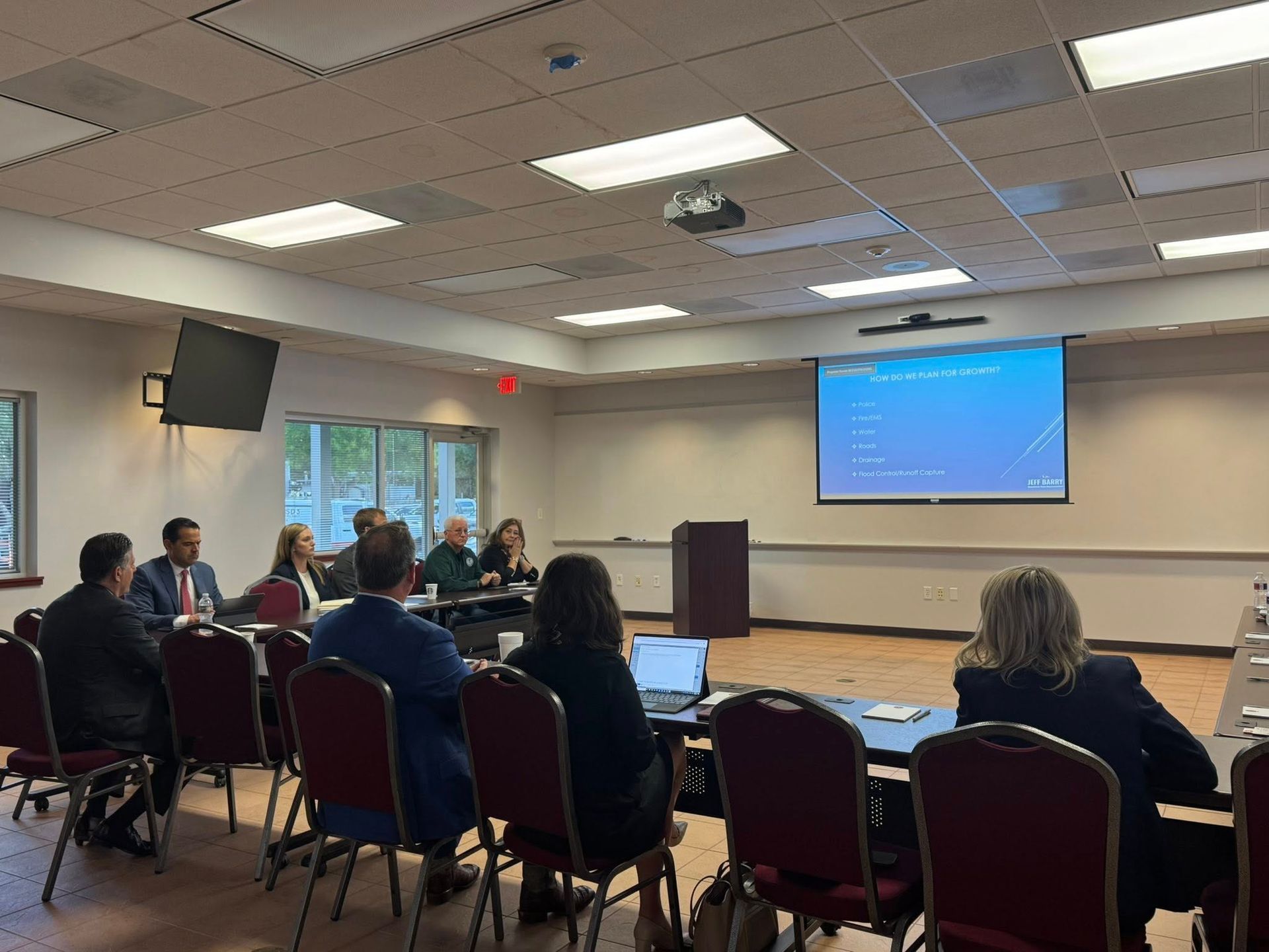 A group of people sit at conference tables in a brightly lit room, facing a projection screen during a presentation.