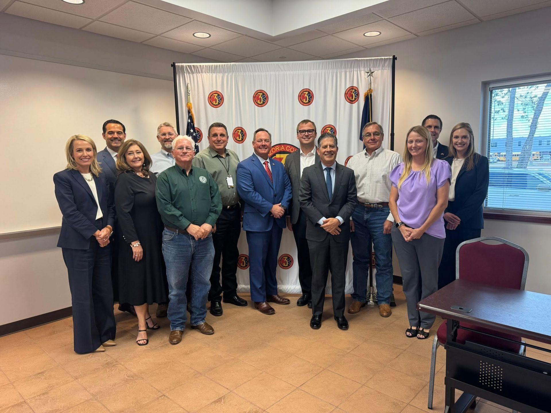 A group of thirteen people stand in two rows smiling for a photo in front of a white curtain with red circular emblems.