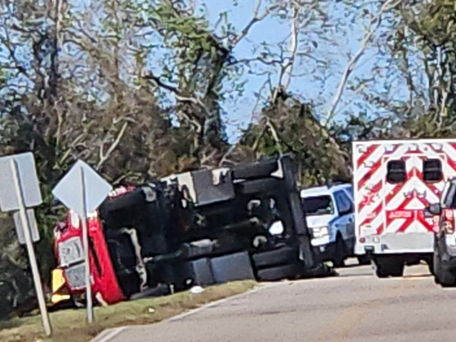 Red fire tanker truck overturned on side of road with emergency vehicles nearby, trees and blue sky background.