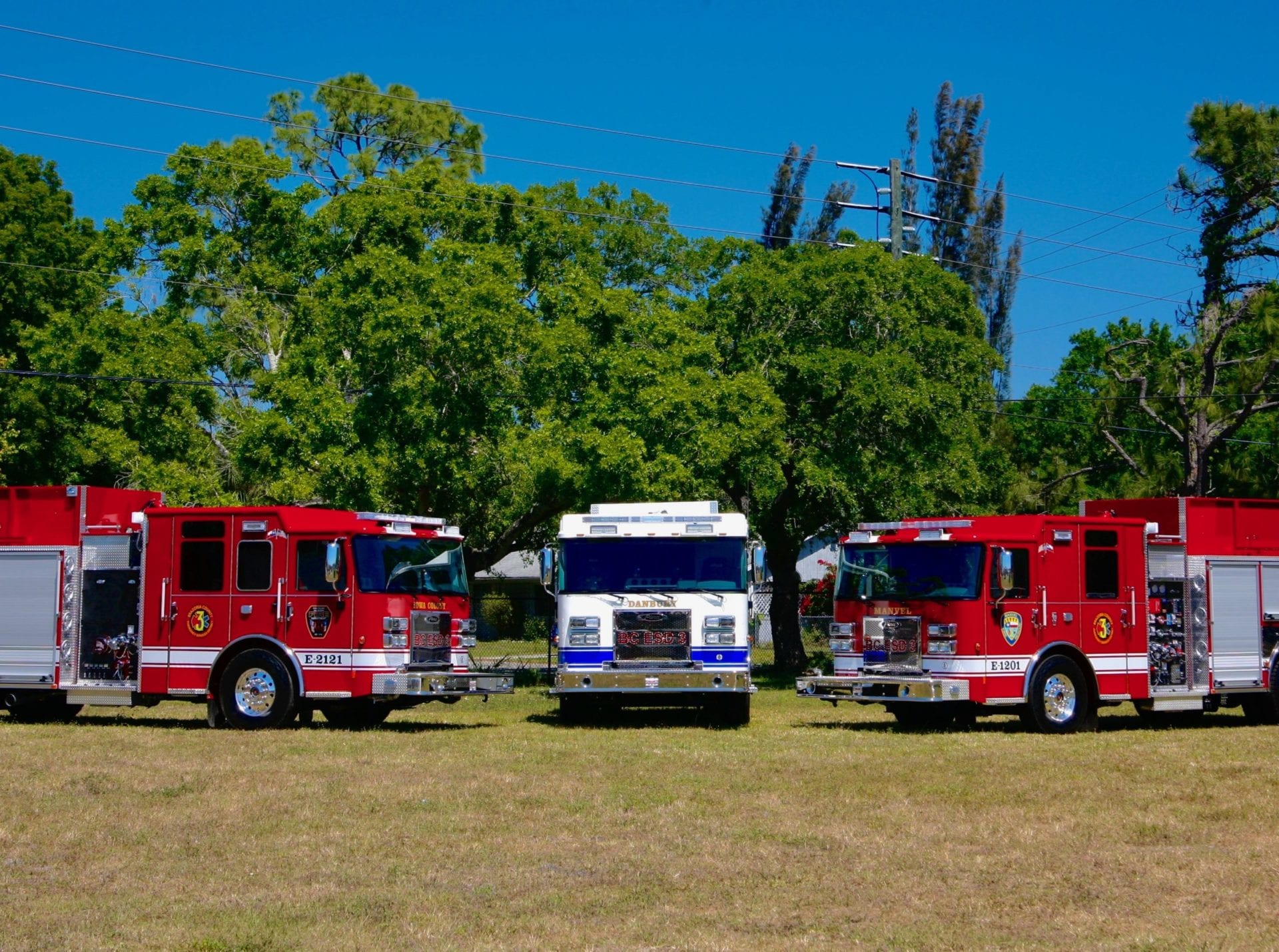 Three fire trucks are parked in a field with trees in the background