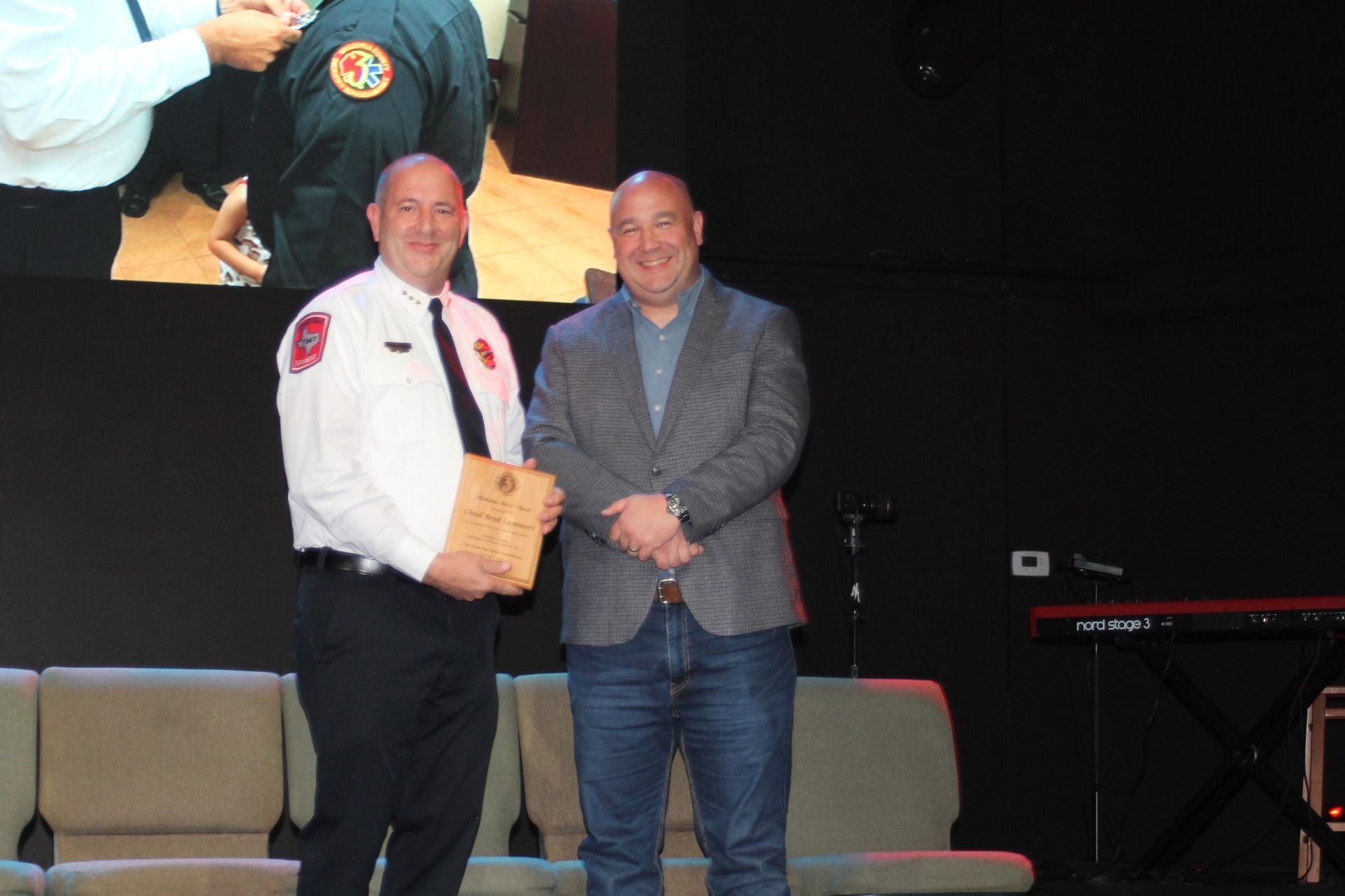 Two men on stage; one in an EMS uniform  holds a plaque, the other in a blazer smiles. They stand in front of a dark backdrop.