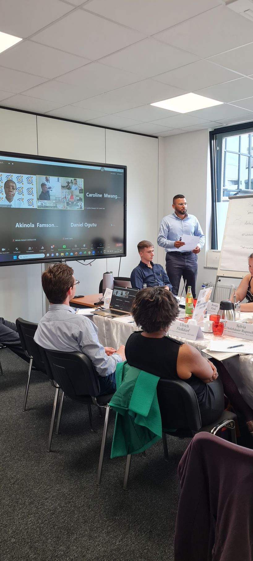 A group of people are sitting at tables in a room while a man stands in front of a large screen.