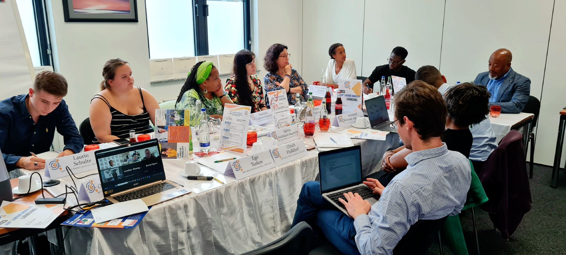 A group of people are sitting around a long table with laptops.