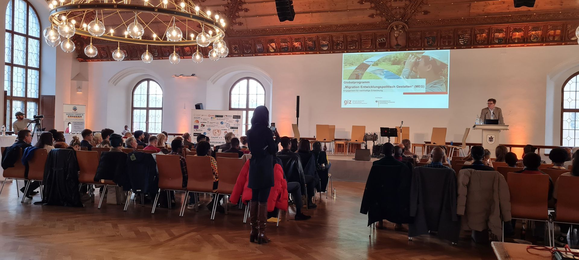 A woman is giving a presentation in front of a large group of people in a large room.