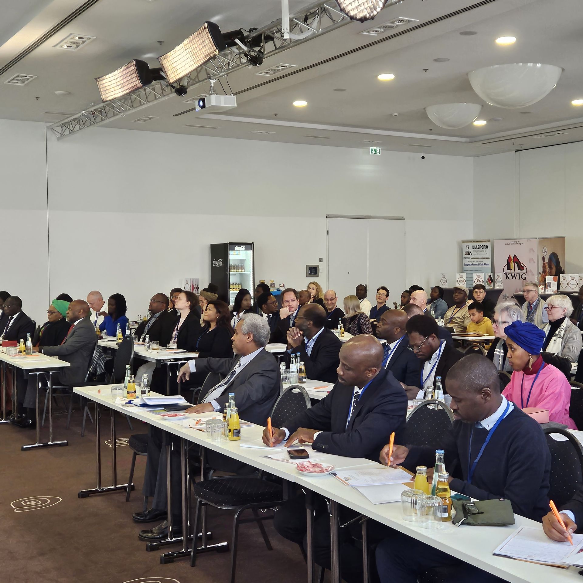 A large group of people are sitting at long tables in a conference room