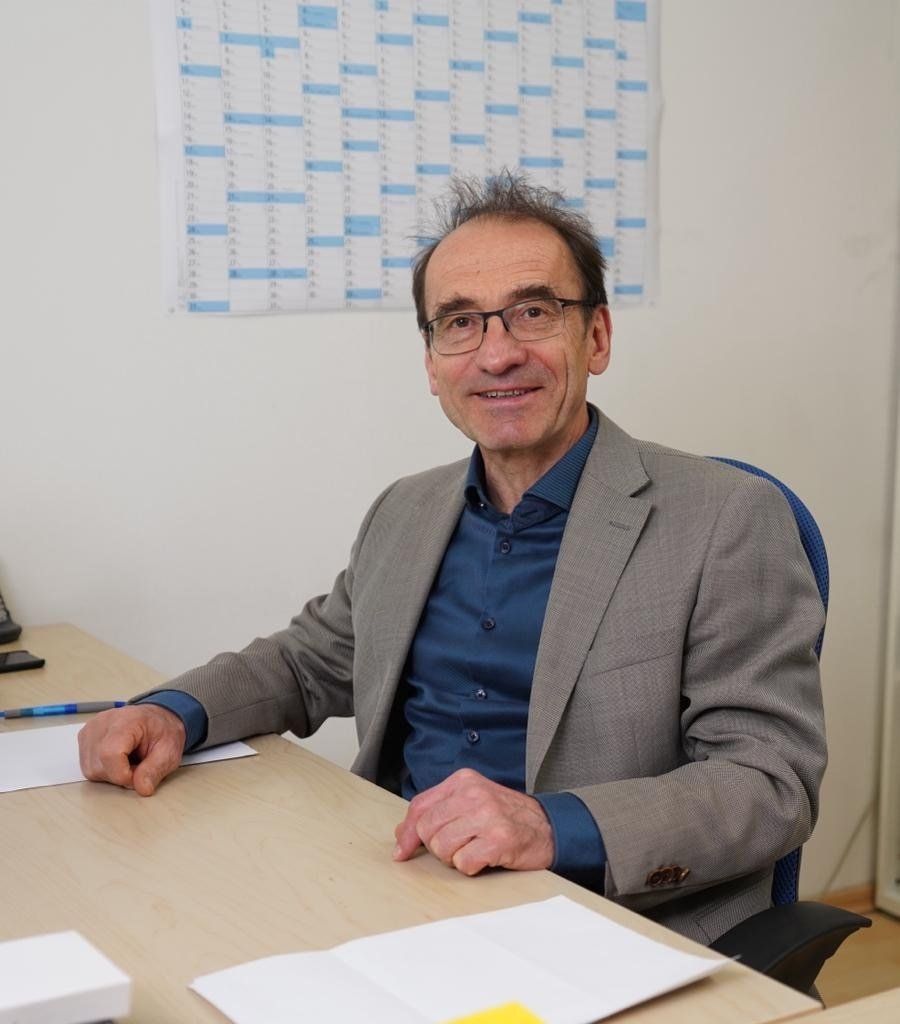 Ein Mann im Anzug sitzt an einem Schreibtisch, hinter ihm hängt ein Kalender an der Wand