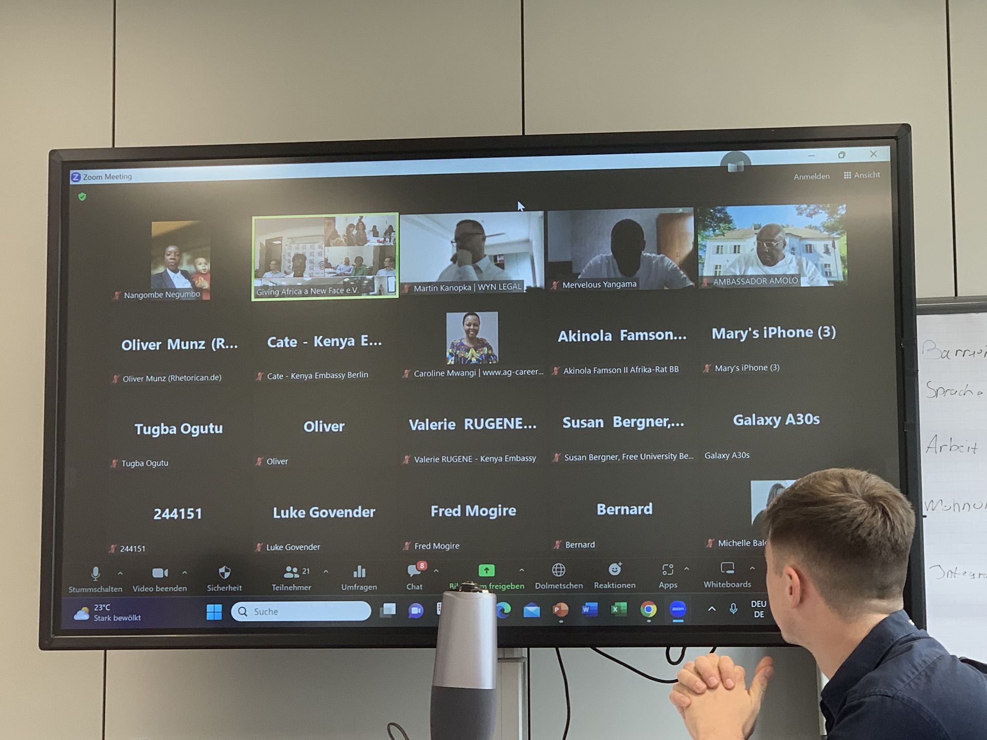A man is sitting in front of a large screen with a group of people on it.