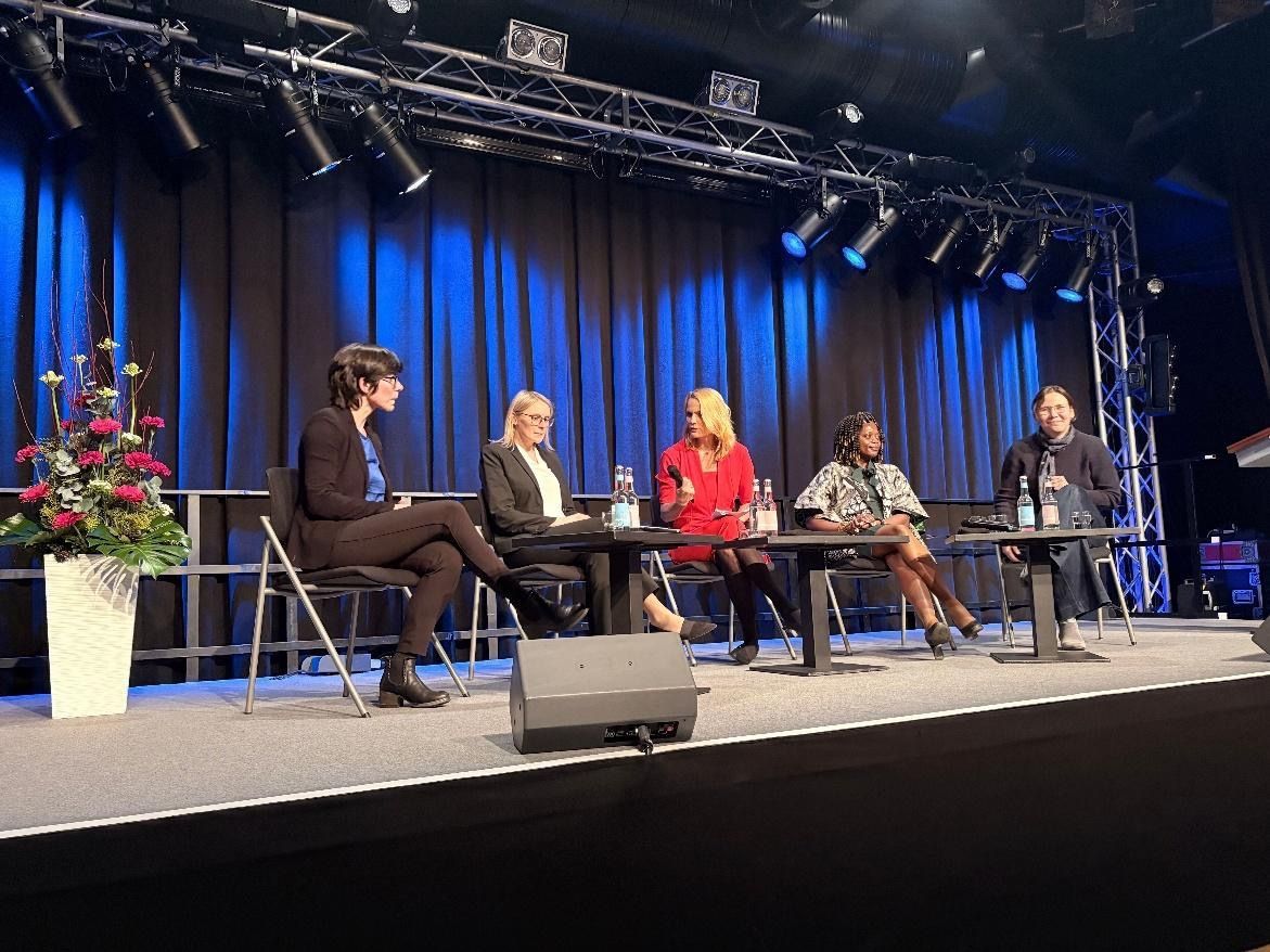 Panel discussion on a stage with five people seated at a table. Blue backdrop, stage lights, and floral arrangement.