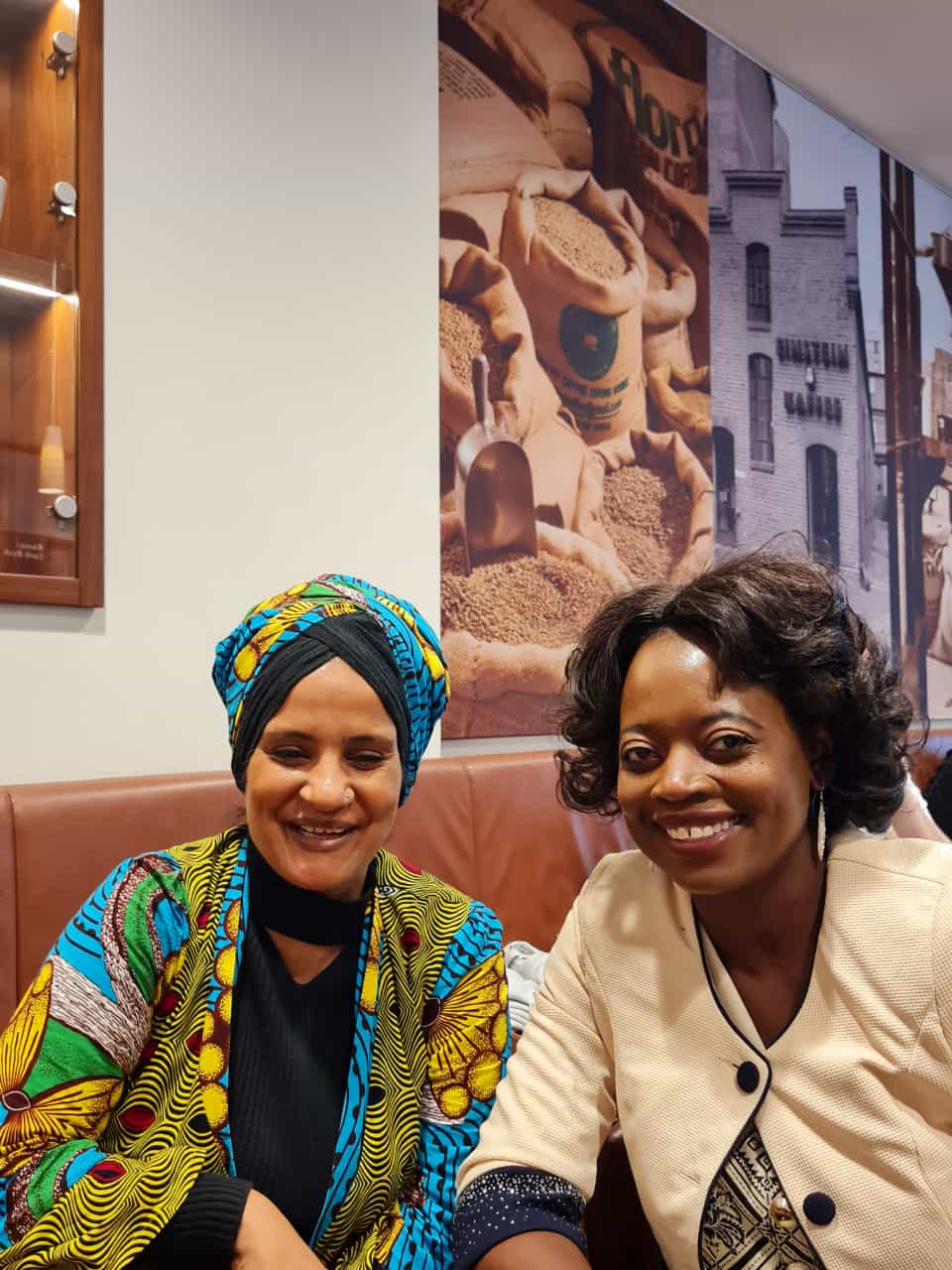 Two women are sitting next to each other in front of a wall with a picture of a coffee shop on it