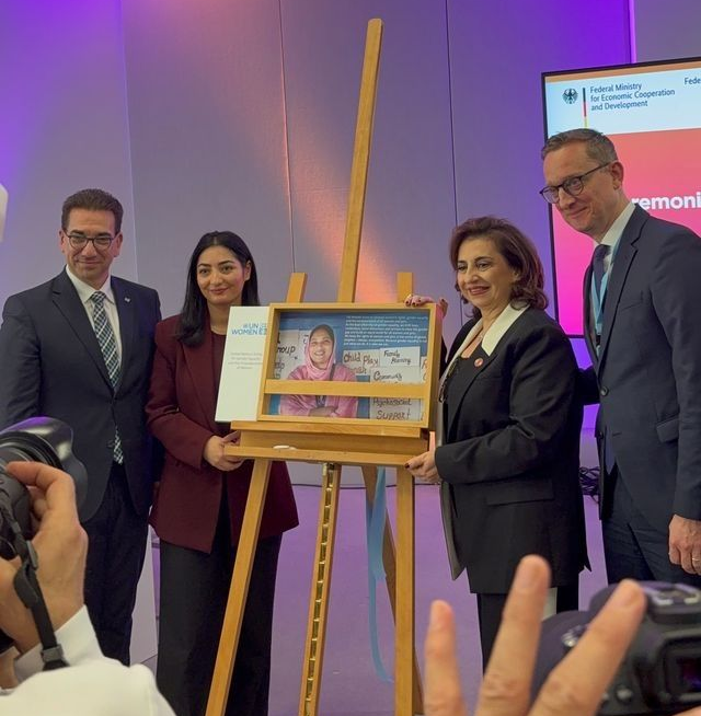 Four people unveil a framed picture on an easel. Formal setting with event signage visible.
