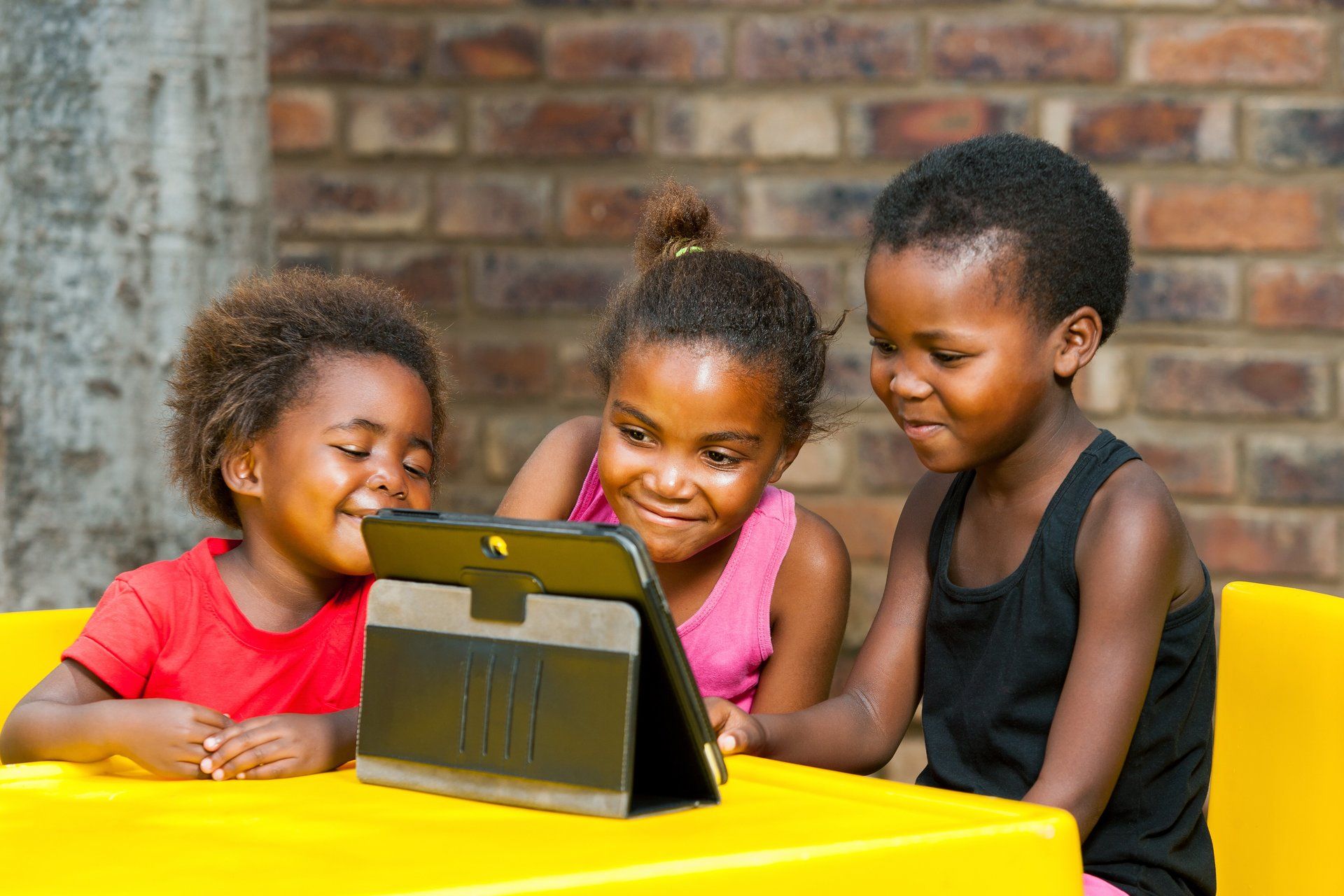 Three children are sitting at a table looking at a tablet.