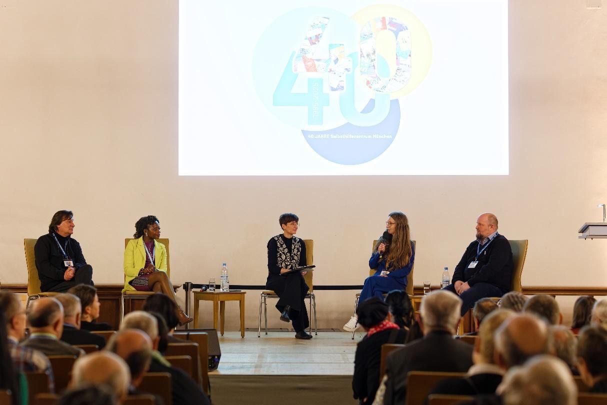 Panel discussion on a stage with five people in front of an audience, projected logo in background.