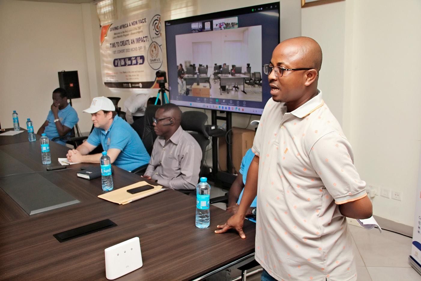 Meeting room with four people seated at a long table and one man standing near a screen.