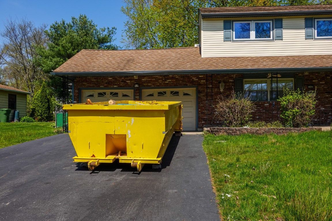 Yellow dumpster on a residential driveway in front of a house with a brown roof and a green lawn.