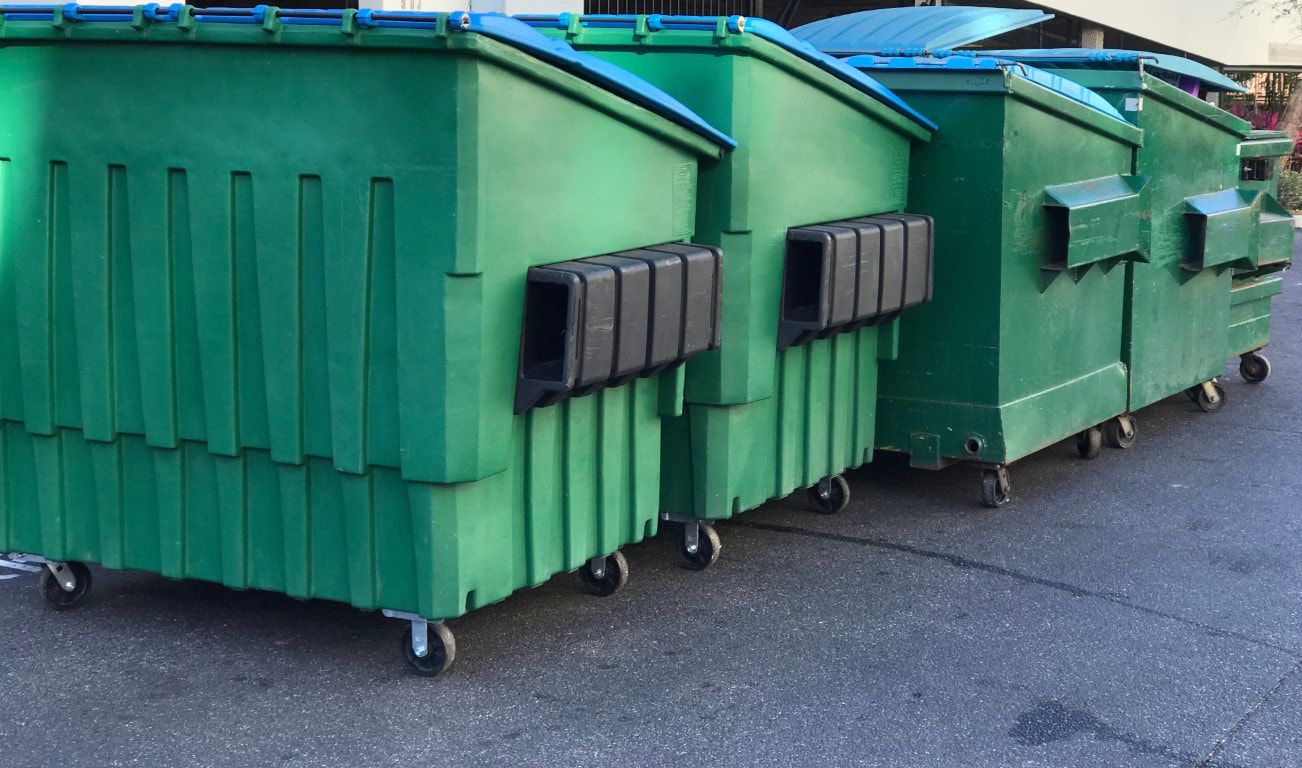 Three green trash dumpsters with blue lids and black openings, lined up on a paved surface.
