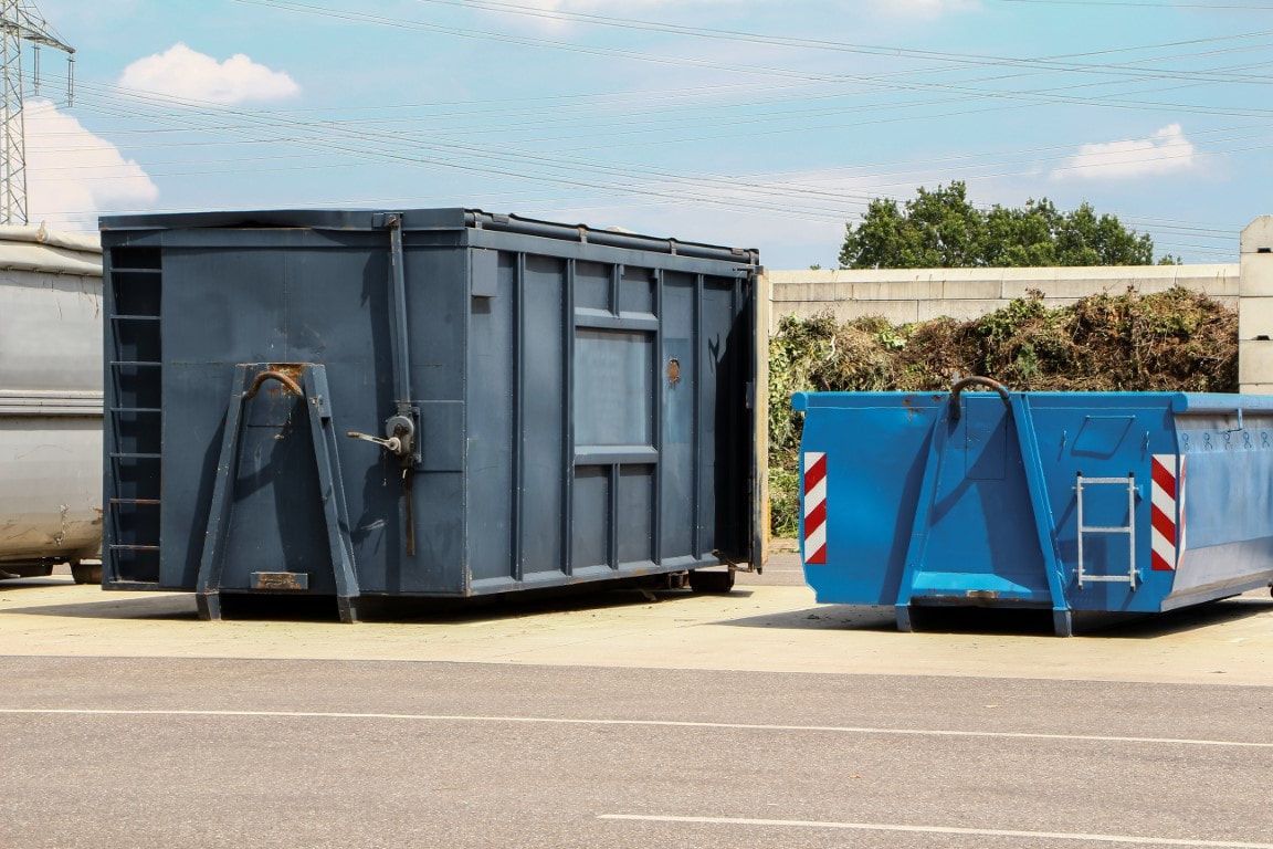 Blue dumpsters at a waste management facility, parked on a paved lot on a sunny day.