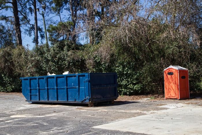 Blue dumpster next to an orange portable toilet, both on a paved area with trees in the background.