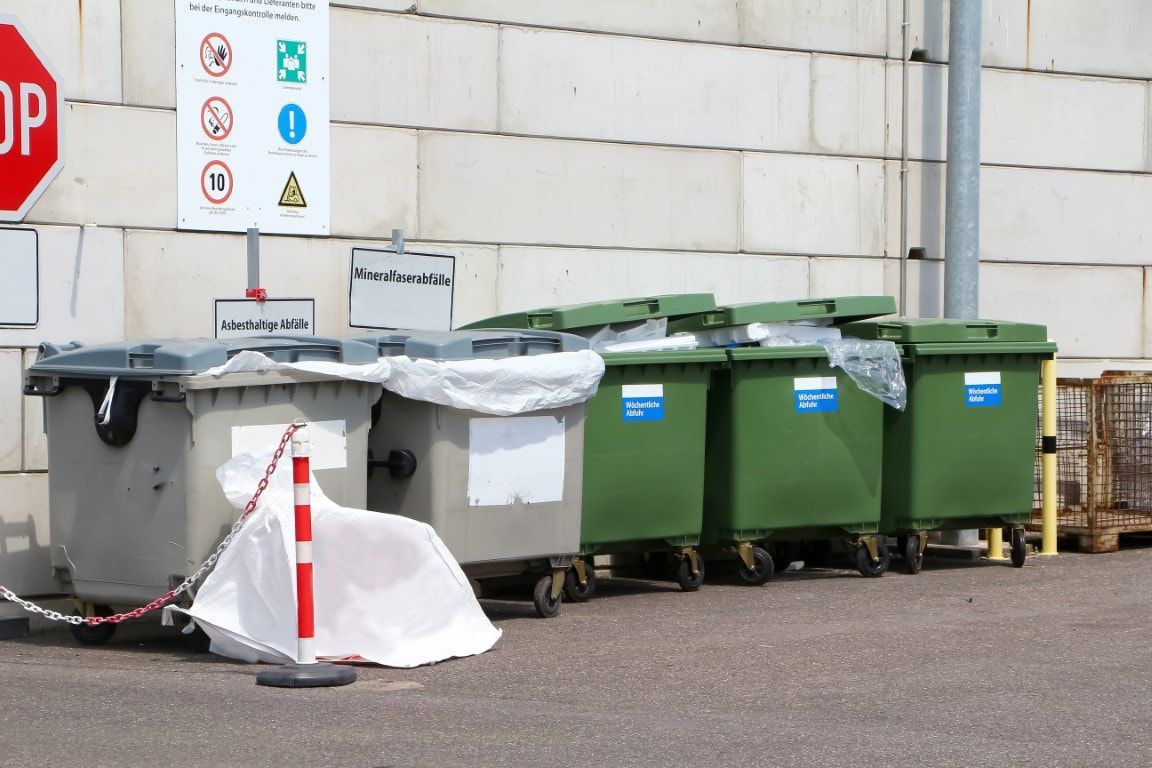 Row of dumpsters in a recycling area; two gray bins, three green bins, with signs and barrier.