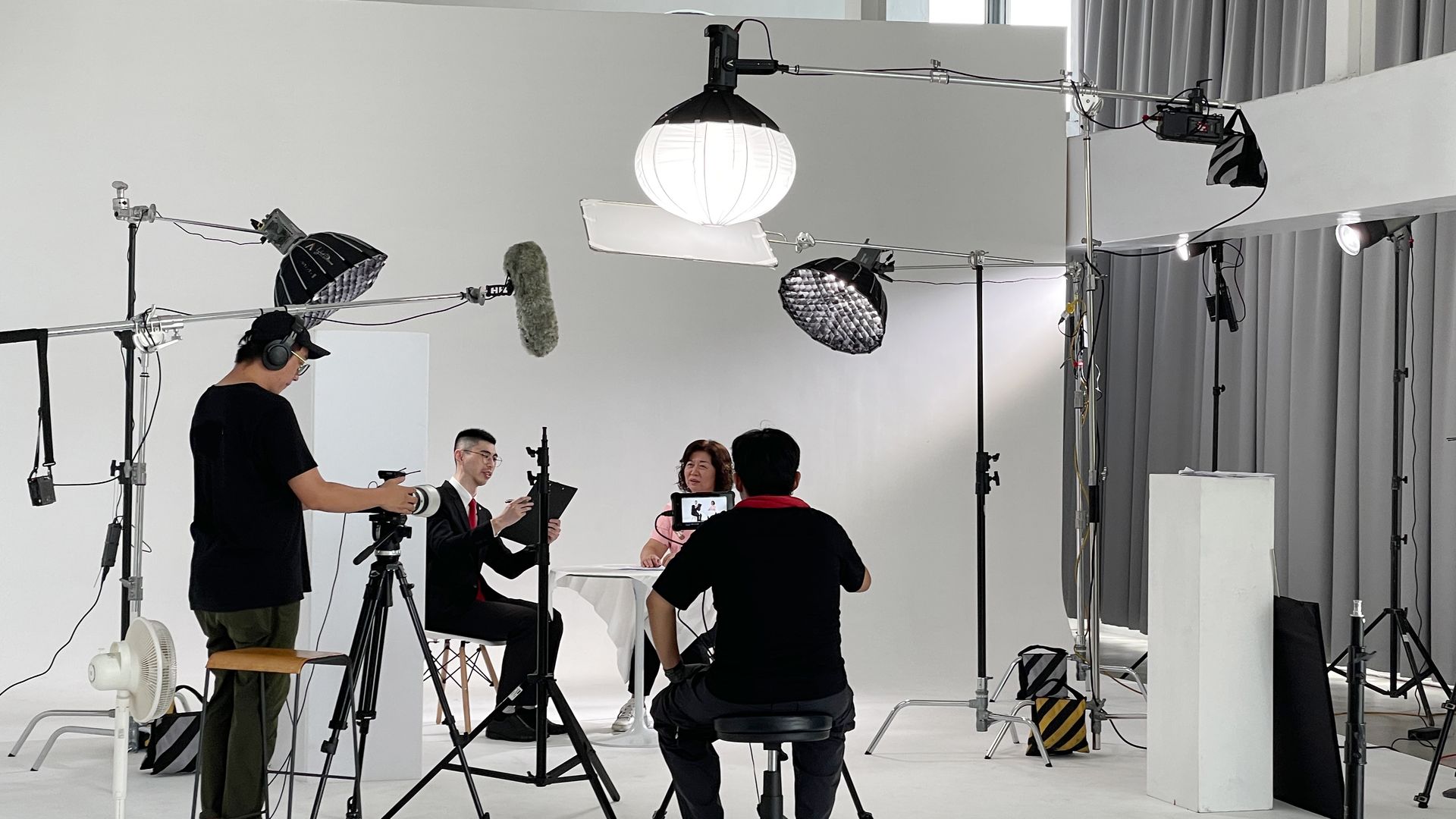 攝影棚內燈光充足，正在拍攝兩人。兩名工作人員操作攝影機和設備。