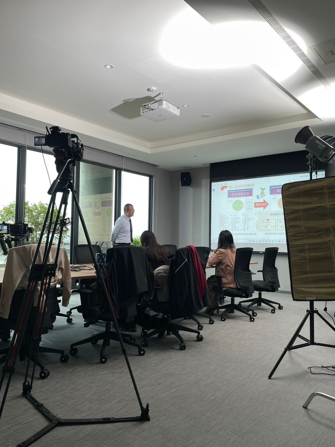 一名男子在會議室裡做演示，會議室裡有一塊投影螢幕。兩台攝影機架在三腳架上。與會者已就座。