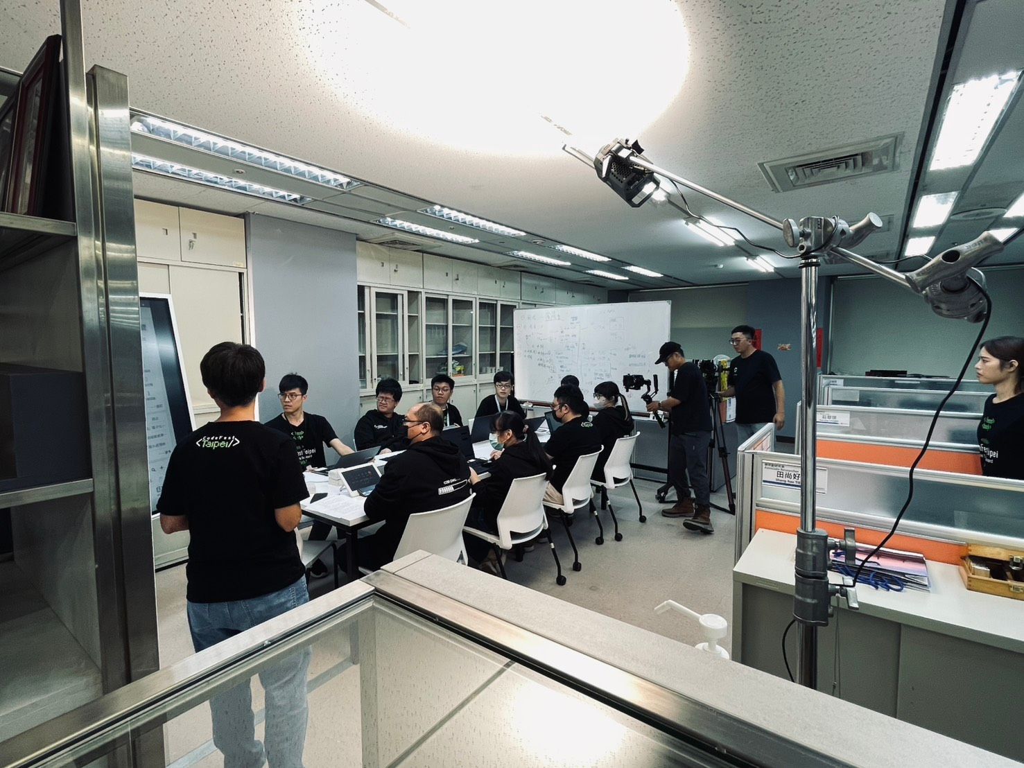 一群人聚集在一間光線充足的辦公室裡，面前有一塊白板，圍坐在一張桌子旁；前面的人正在向他們講話。