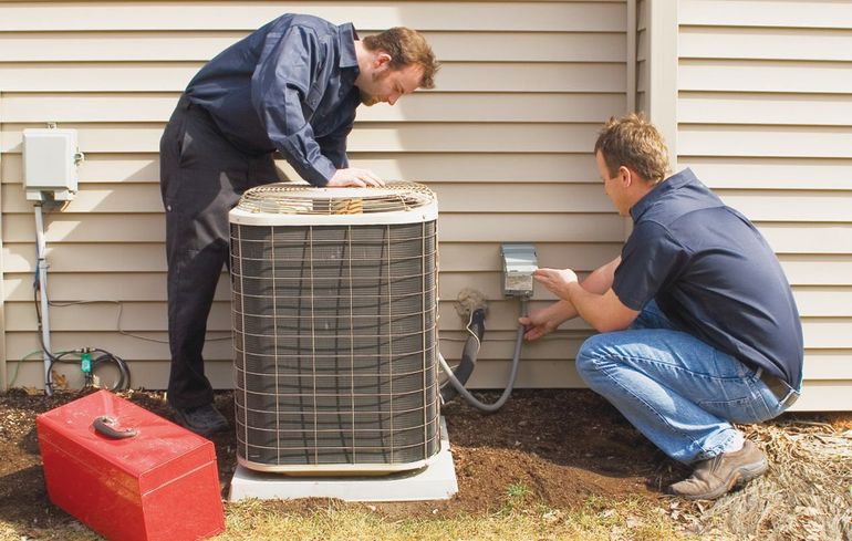 Two technicians inspect an outdoor air conditioner beside a house, with tools and a red toolbox nearby
