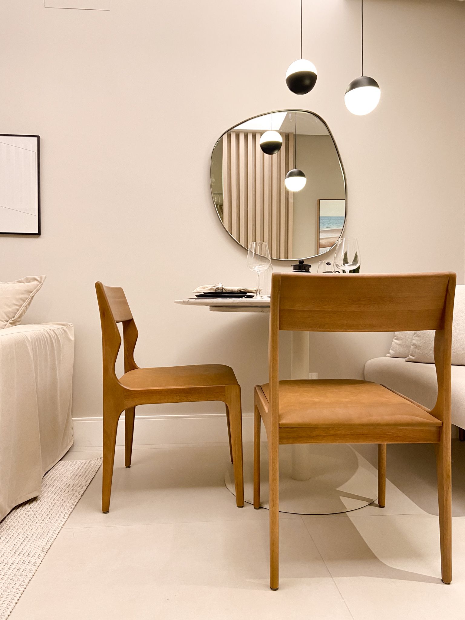 Duas cadeiras de madeira ficam de frente para uma pequena mesa sob luminárias pendentes, com um espelho exclusivo e uma parede bege ao fundo.