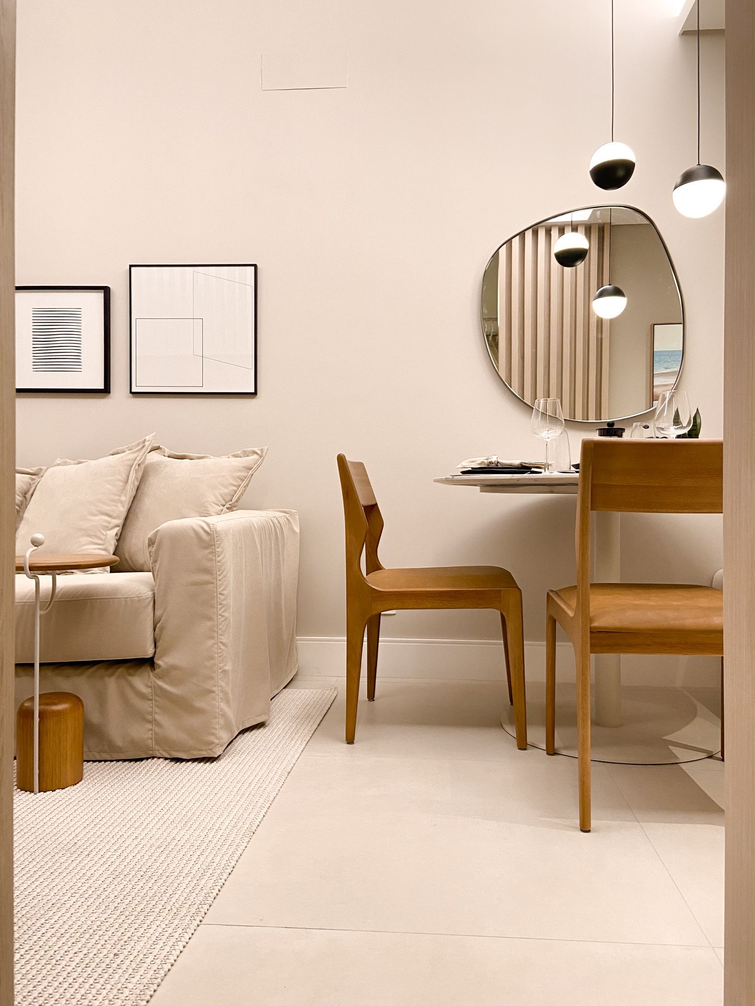 Sala de estar bege com sofá, obra de arte, mesa, cadeiras, espelho e luminárias pendentes.
